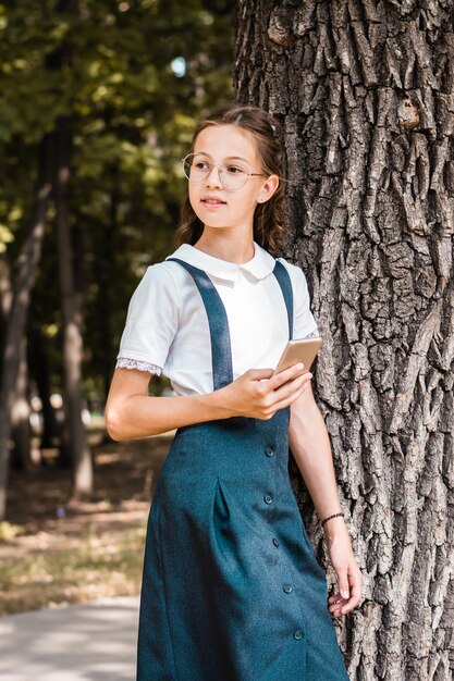 Foto ein schulmädchen mit brille und smartphone in der hand steht in der nähe eines baumes im park