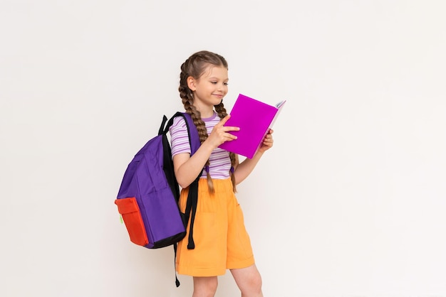 Ein Schulmädchen mit einem Schulranzen hinter ihr liest ein Buch auf weißem, isoliertem Hintergrund Sommerferienkurse