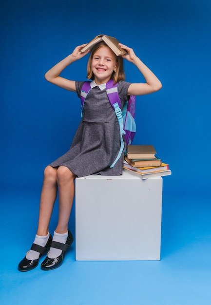 Ein Schulmädchen sitzt auf einem weißen Würfel und liest ein Buch, das auf blauem Hintergrund isoliert ist, zurück zur Schule