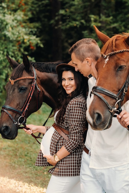Ein schwangeres Mädchen in einem Hut und ihr Mann in weißen Kleidern stehen neben Pferden im Wald in der Natur. Stylish schwangere Frau mit einem Mann mit Pferden. Familie.