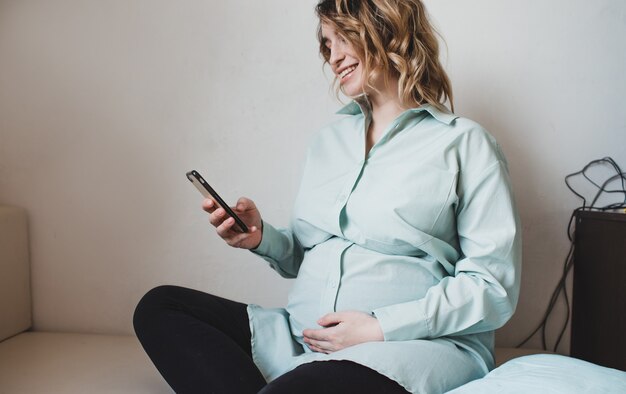Ein schwangeres Mädchen in einem türkisfarbenen Hemd hält ein Telefon in ihren Händen. Warten auf eine Schwangerschaft