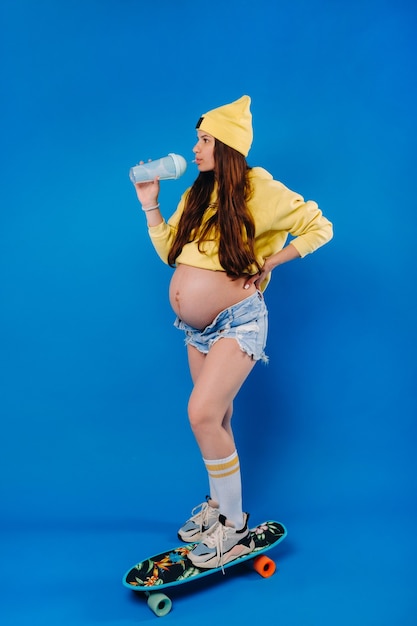 Ein schwangeres Mädchen in gelber Kleidung mit einem Glas Saft fährt ein Skateboard auf blauem Hintergrund