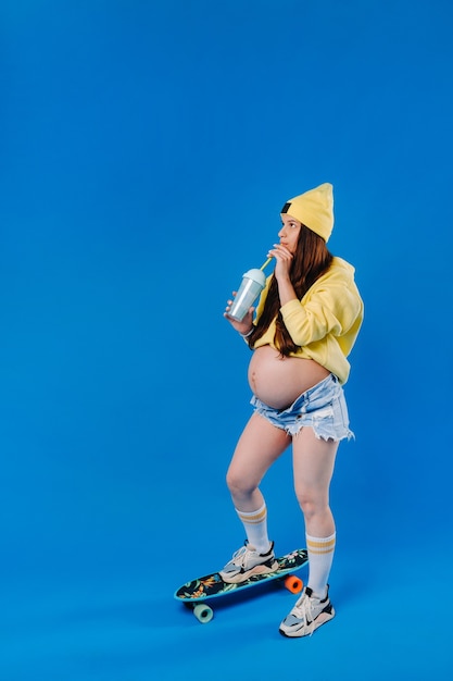 Ein schwangeres Mädchen in gelber Kleidung mit einem Glas Saft fährt ein Skateboard auf blauem Hintergrund.