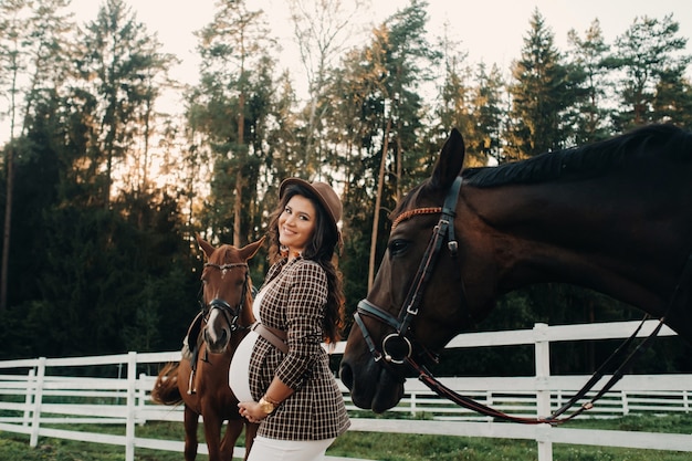 Ein schwangeres Mädchen mit einem dicken Bauch in einem Hut neben Pferden nahe einer Koppel in der Natur. Stylish schwangere Frau in einem braunen Kleid mit Pferden.