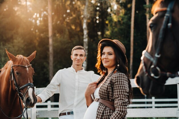 Ein schwangeres Mädchen mit Hut und ihr Mann in weißer Kleidung stehen neben den Pferden in der Nähe des Pferdes. Stylish schwangere Frau mit einem Mann mit Pferden. Familie.