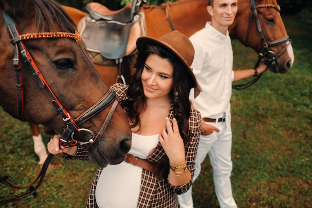 Ein schwangeres Mädchen mit Hut und ihr Mann in weißer Kleidung stehen neben Pferden im Wald in der Natur. Stilvolle schwangere Frau mit einem Mann mit Pferden. Familie.