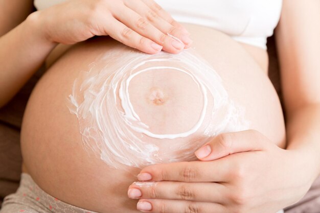 Ein schwangeres Mädchen sitzt zu Hause auf dem Bett und schmiert eine Creme gegen Dehnungsstreifen auf den Bauch Schwangerschaftsmutterschaftsvorbereitung und Erwartungskonzept