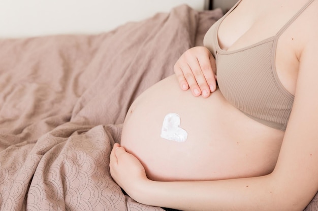 Ein schwangeres Mädchen sitzt zu Hause auf dem Bett und schmiert Herz eine Creme gegen Dehnungsstreifen auf den Bauch Schwangerschaft Mutterschaftsvorbereitung und Erwartungskonzept