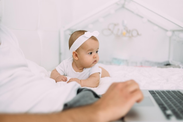 Ein sechs Monate altes Baby liegt auf einem weißen Bett und sieht Papa beim Lernen zu, während es neben einem Laptop sitzt