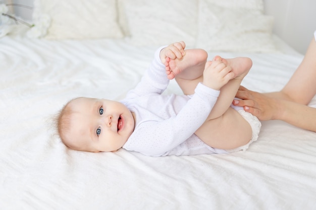 Ein sechs Monate altes Baby mit den Händen seiner Mutter auf einem weißen Baumwollbett zu Hause lächelnd oder lachend or