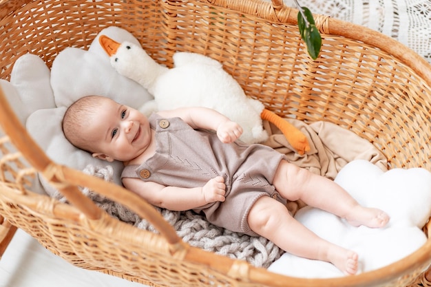 Ein sechs Monate altes Mädchen im Kinderzimmer in einer Korbwiege zu Hause mit einem Stofftier, das lächelt oder lacht. Ein lächelndes kleines Kind mit einem Spielzeug in den Händen auf dem Bett im Schlafzimmer