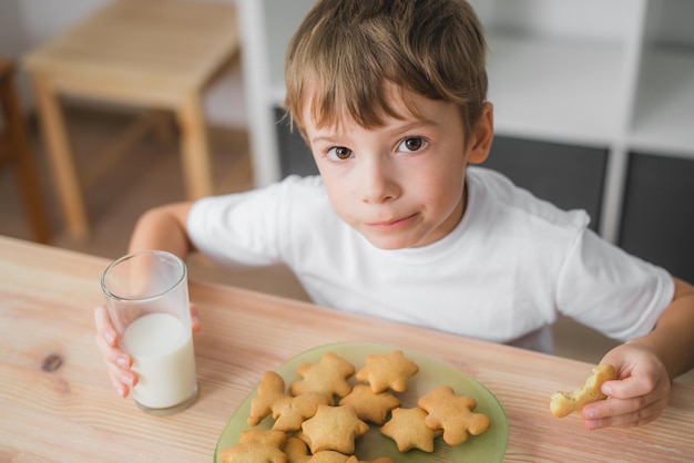 Ein sechsjähriger Junge frühstückt mit Keksen und Milch, sitzt am Tisch und schaut in die Kamera. Er hat braune Augen und hellbraune Haare und trägt ein weißes T-Shirt. Hausmannskost