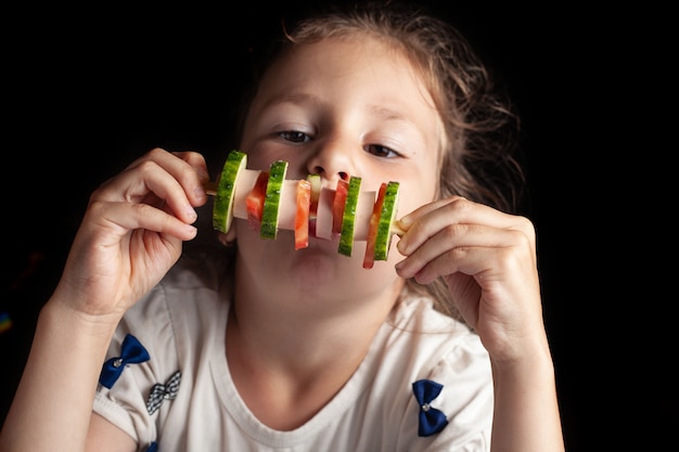 Ein sechsjähriges Mädchen isst ein ungewöhnliches Sandwich am Stiel. Gurke, Tomate, Wurst