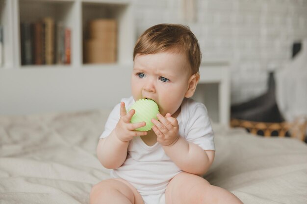 Ein sechsmonatiges Baby, das den Ball mit den Händen hält und auf dem Bett sitzt, reißt die Zähne.