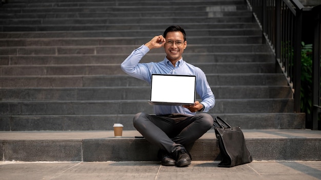 Foto ein selbstbewusster asiatischer geschäftsmann sitzt auf einer außentreppe und hält einen laptop mit leerem bildschirm