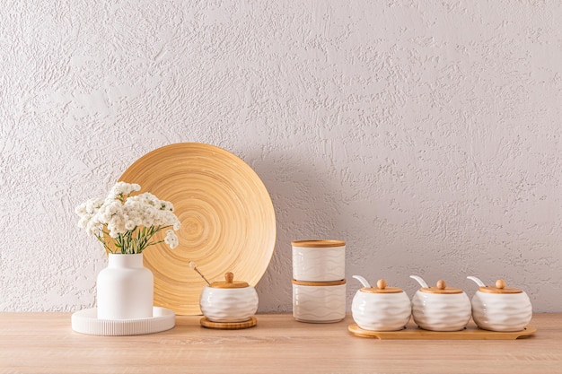 Ein Set aus weißen Keramikgläsern mit Bambusdeckeln zur Aufbewahrung von Massenprodukten und Gewürzen auf einer Holzarbeitsplatte mit weißen Blumen in einer Vase