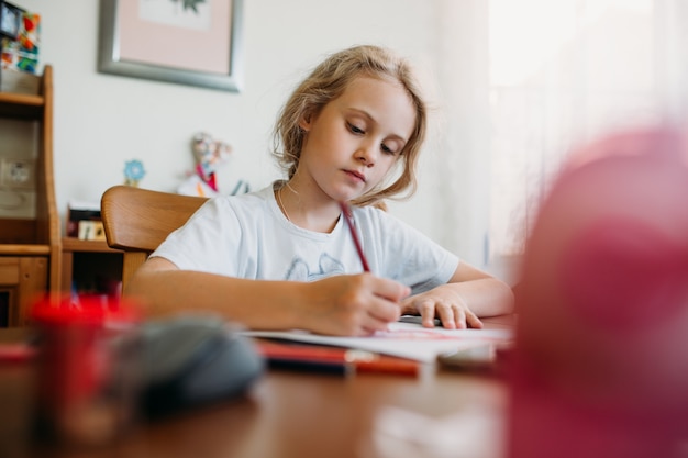 Ein siebenjähriges Mädchen sitzt zu Hause an einem Tisch und schreibt in ein Notizbuch, erledigt eine Lernaufgabe oder wiederholt Lektionen.