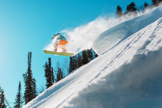 Ein Snowboarder springt von einem hohen schneebedeckten Sprungbrett in einem Wald gegen einen blauen Himmel. Extremsportarten