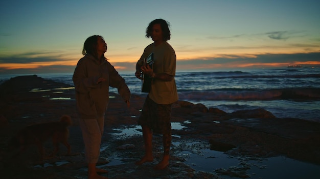 Ein sorgloses Paar tanzt zusammen am Abend am Strand, ein Mann spielt Gitarre und küsst ein Mädchen.
