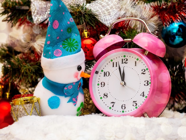 Ein Spielzeug-Schneemann in der Nähe der Uhr, der die Annäherung von 12 Stunden, dem neuen Jahr, anzeigt. Schneemann, Uhr neben dem Weihnachtsbaum_