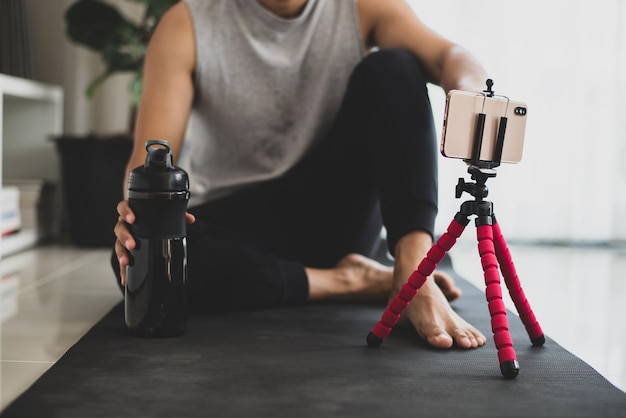 Ein Sportler sitzt auf der Trainingsmatte und hält einen Proteinshake in einer schwarzen Flasche und nimmt mit dem Smartphone Online-Yoga-Kurse über einen Videoanruf auf dem Laptop im heimischen Wohnzimmer