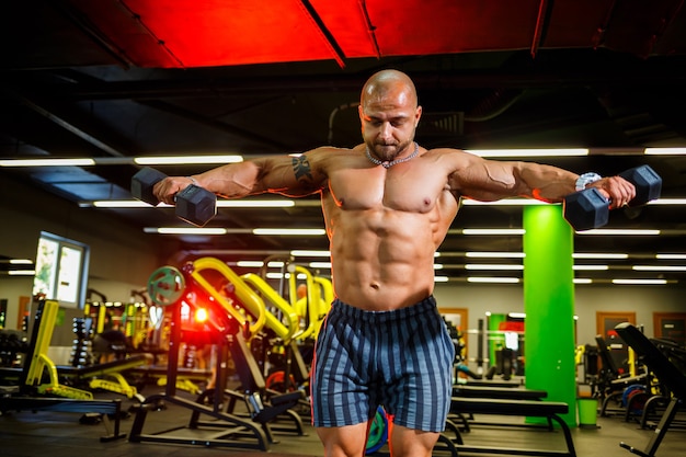 Ein starker, erwachsener, fitter, muskulöser männlicher Trainer posiert für ein Fotoshooting in einer Sporthalle im Scheinwerferlicht in Sportkleidung, demonstriert seine Muskeln und hält souverän Hanteln.