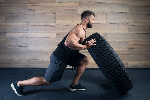 Ein starker Mann in einem schwarzen Trägershirt und grauen Shorts mit Bart versucht, in einem Fitnessstudio einen Reifen zu schieben