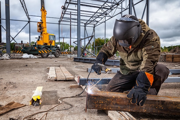 Ein starker mann ist ein schweißer in brauner uniform schweißmaske und schweißerleder ein metallprodukt wird mit einem lichtbogenschweißgerät auf der baustelle geschweißt blaue funken fliegen zu den seiten