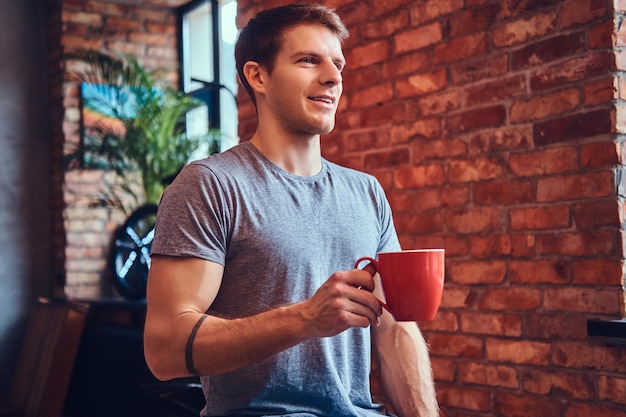 Ein stilvoller, attraktiver Mann in Jeans und T-Shirt, der auf einem Holzstuhl sitzt und eine Tasse Kaffee hält. Wegschauen.