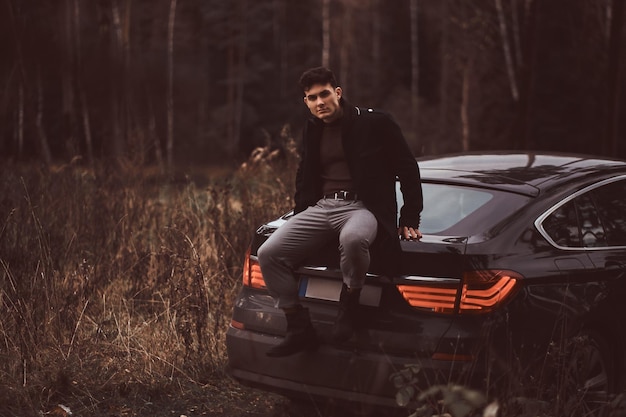 Ein stilvoller junger Mann mit schwarzem Mantel, der abends im Herbstwald auf dem Kofferraum eines Autos sitzt.