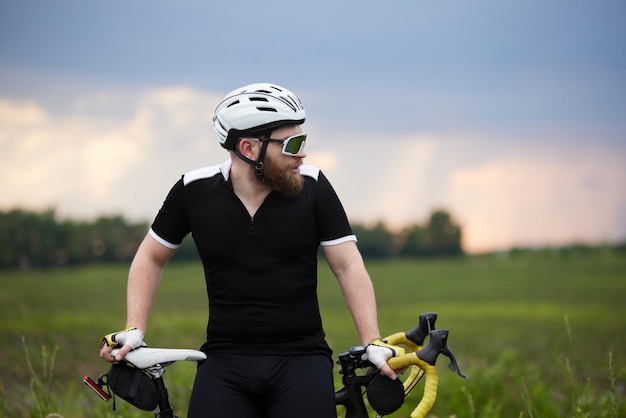 Ein stilvoller, sportlicher, bärtiger Mann in Sportbekleidung und Helm sitzt auf einem Fahrrad am Straßenrand auf einem Feld in der Kälte