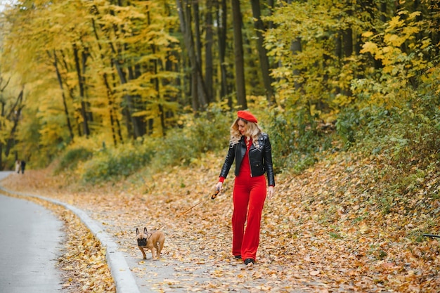 Ein stilvolles junges Mädchen mit langen hellen Haaren in sonniger Brille geht im Frühjahr im Herbst mit einem kleinen mittleren Hündchen einen Mops von der französischen Bulldogge in einem Park spazieren