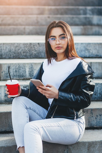 Ein stilvolles Mädchen mit Brille und weißem T-Shirt benutzt Smartphone und trinkt Kaffee