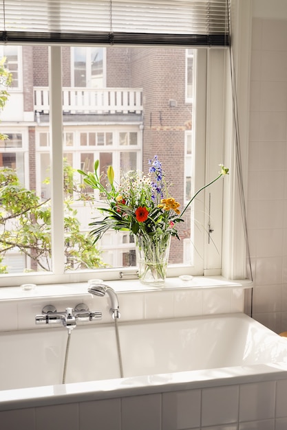 Ein Strauß frischer Frühlingsblumen in einer Vase, die an einem sonnigen Tag auf einem Fensterbrett in einem großen hellweißen Badezimmer steht. Gemütliche Inneneinrichtung für Ihr Zuhause.