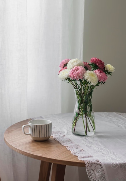 Ein Strauß Herbstblumen mit Astern in einer Glasvase und einer Keramiktasse auf einem runden Tisch im Wohnzimmer