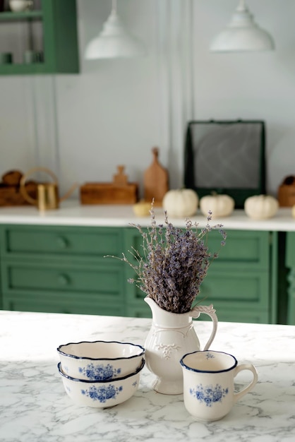 Ein Strauß Lavendel und eine Reihe weißer Gerichte, ein Milchkännchen aus Porzellan, eine Kaffeetasse und Schalen auf einer weißen Arbeitsplatte vor einer grünen Küche. Weicher selektiver Fokus