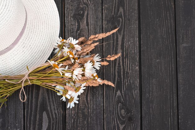 Ein Strauß weißer Gänseblümchen, Blumen mit trockenen Ohren in einer Kraftpapiervase mit einem Hut auf einem schwarzen hölzernen Hintergrund in einem rustikalen Stil.