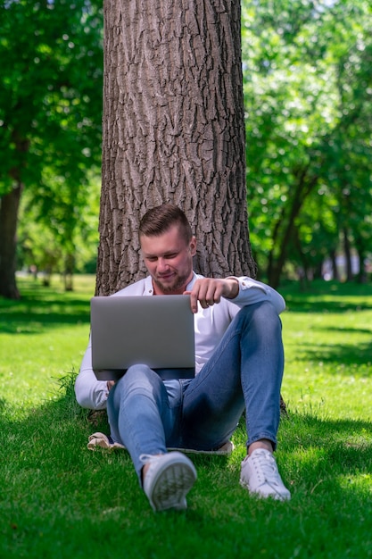 Ein Student, der ein Video in einem Laptop sieht, der an einem sonnigen Tag im Gras neben einem Baum in einem Landschaftspark sitzt