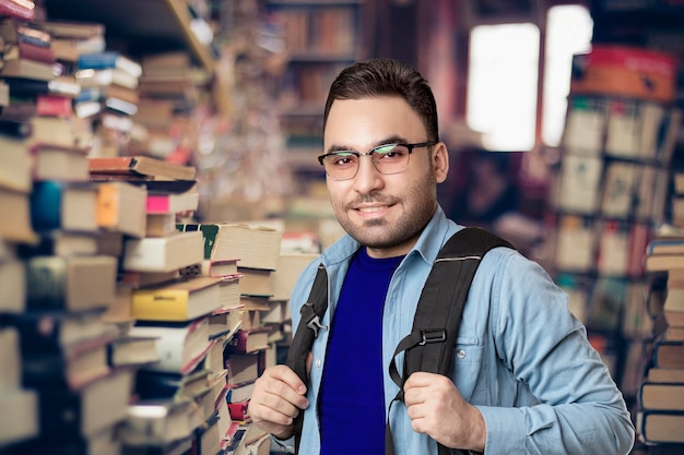 Ein Student, der in einer Bibliothek steht, einen Rucksack trägt, in die Kamera schaut und lächelt