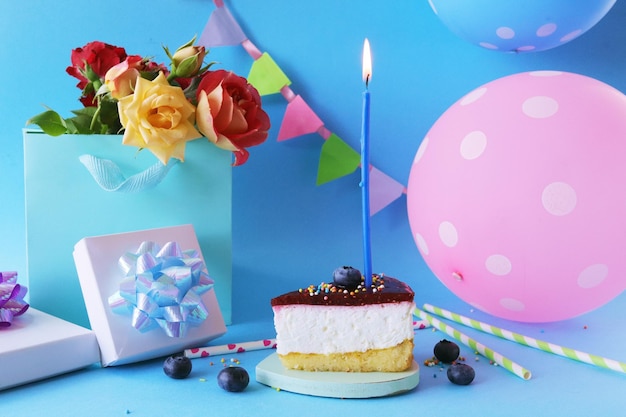 Ein Stück Beerenkuchen mit brennender Kerze, ein Strauß frischer Rosen, Geschenke, Luftballons