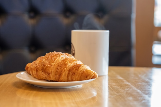 Ein Stück Croissant auf einem weißen Teller mit einer Tasse heißen Kaffees auf Holztisch im Café