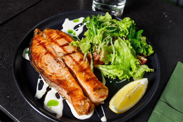 Ein Stück gebackener Lachs gegrillte Pfefferzitrone und Salz auf einem schwarzen Teller mit Salatblättern auf Steinhintergrund