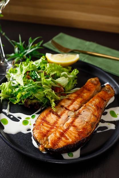 Ein Stück gebackener Lachs gegrillte Pfefferzitrone und Salz auf einem schwarzen Teller mit Salatblättern auf Steinhintergrund