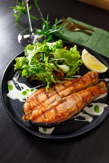 Ein Stück gebackener Lachs gegrillte Pfefferzitrone und Salz auf einem schwarzen Teller mit Salatblättern auf Steinhintergrund