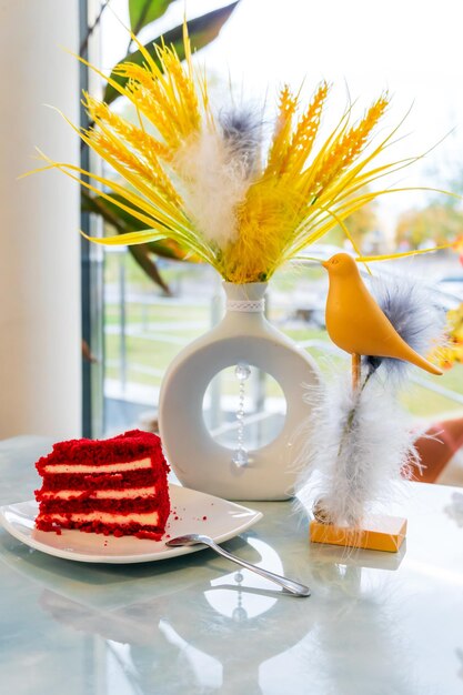 Ein Stück köstlicher Kuchen auf einem Teller und eine Tasse Kaffee auf dem Tisch. Auf dem Tisch im Restaurant Couchtisch neben dem Fenster stehen künstliche gelbe Blumen und ein Spielzeugvogel
