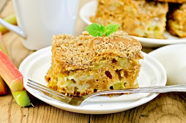 Ein Stück süßer Kuchen mit Rhabarber, Minze, weißer Serviette, Tasse auf dem Hintergrund von Holzbrettern