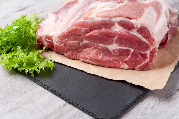 Ein Stück ungekochtes Fleisch vom Schweinenacken auf schwarzem Stometeller mit Zutat auf grauem Tisch. Bereit zum Kochen. Nahaufnahme.