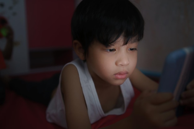 Ein süßer asiatischer Junge, der auf seinen Handys liegt, spielt Smartphone-Nomophobie bei Kindern