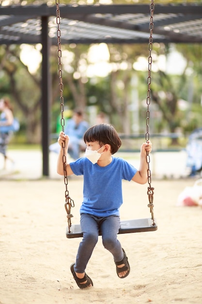Ein süßer asiatischer Junge, der eine Maske trägt, spielt tagsüber im Sommer auf einer Schaukel auf dem Spielplatz Outdoor-Aktivitäten Play Makes Ideas Believe Externes Bildungsporträt