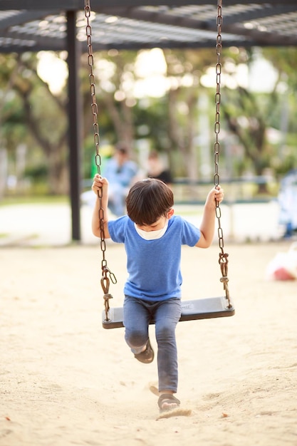 Ein süßer asiatischer Junge, der eine Maske trägt, spielt tagsüber im Sommer auf einer Schaukel auf dem Spielplatz Outdoor-Aktivitäten Play Makes Ideas Believe Externes Bildungsporträt